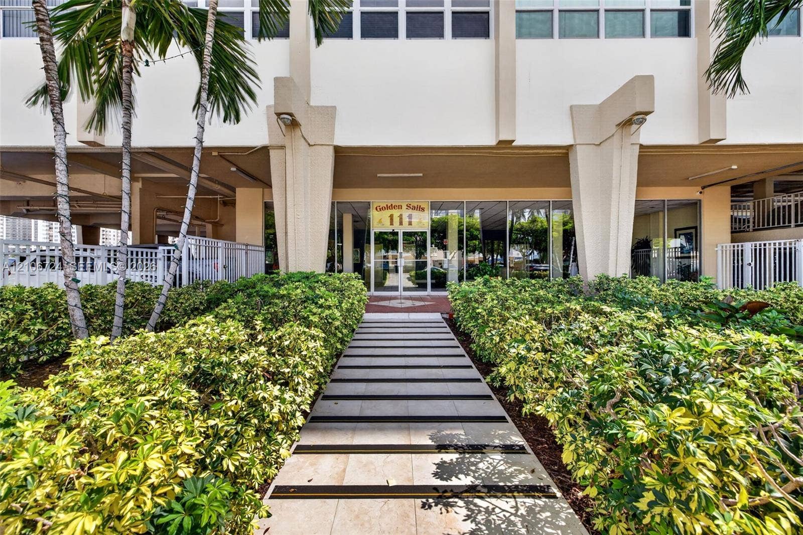 111 Golden Isles Drive, #G12, Hallandale Beach, FL 33009 Building entrance featuring structural pillars, floor-to-ceiling glass paneling, a paved walkway with inlaid strips, and manicured landscaping with palm trees