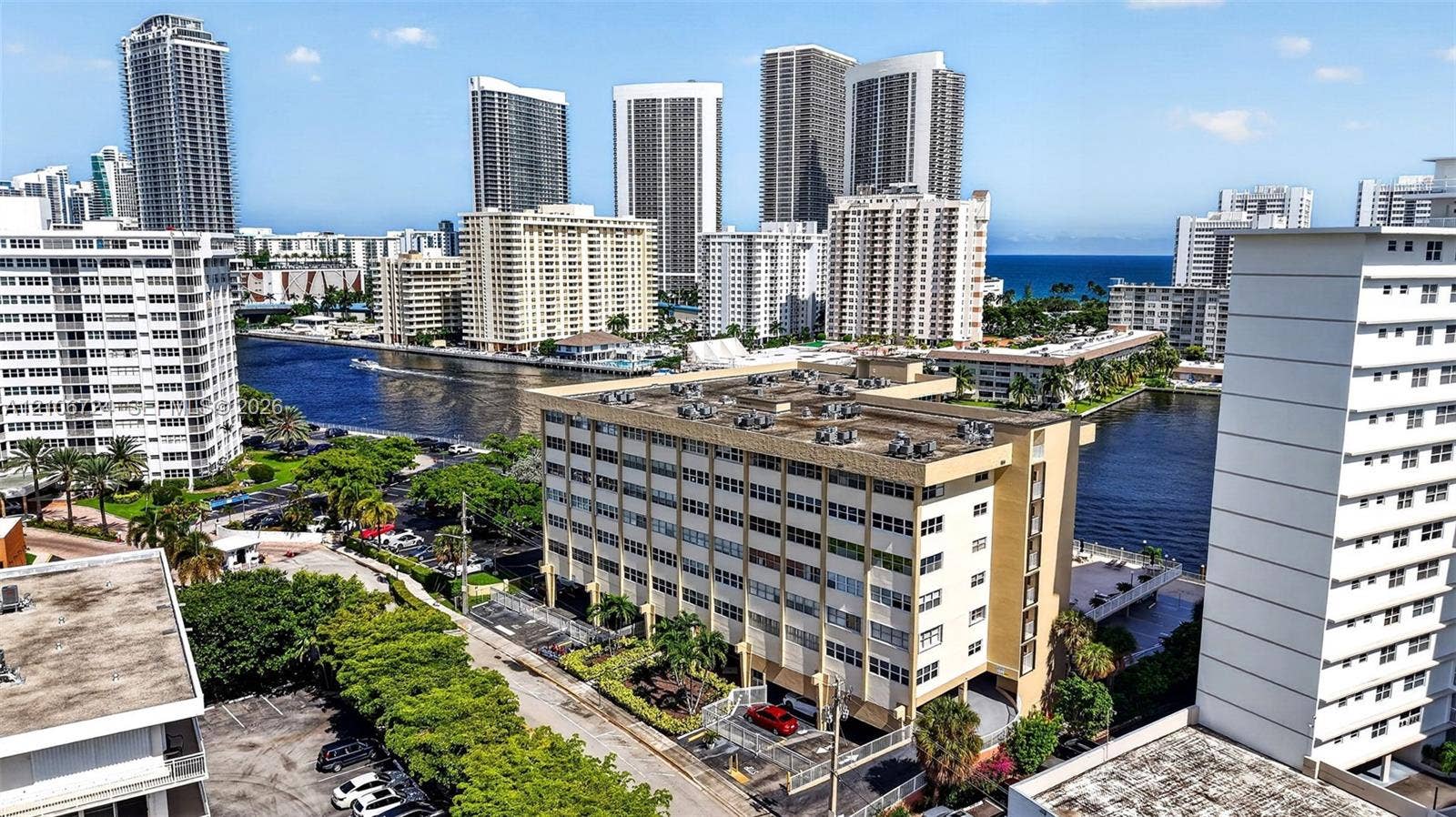 111 Golden Isles Drive, #G12, Hallandale Beach, FL 33009 Mid-rise multi-story building featuring a concrete facade, flat roof with roof-mounted HVAC units, ground-level covered parking, and an urban waterfront location