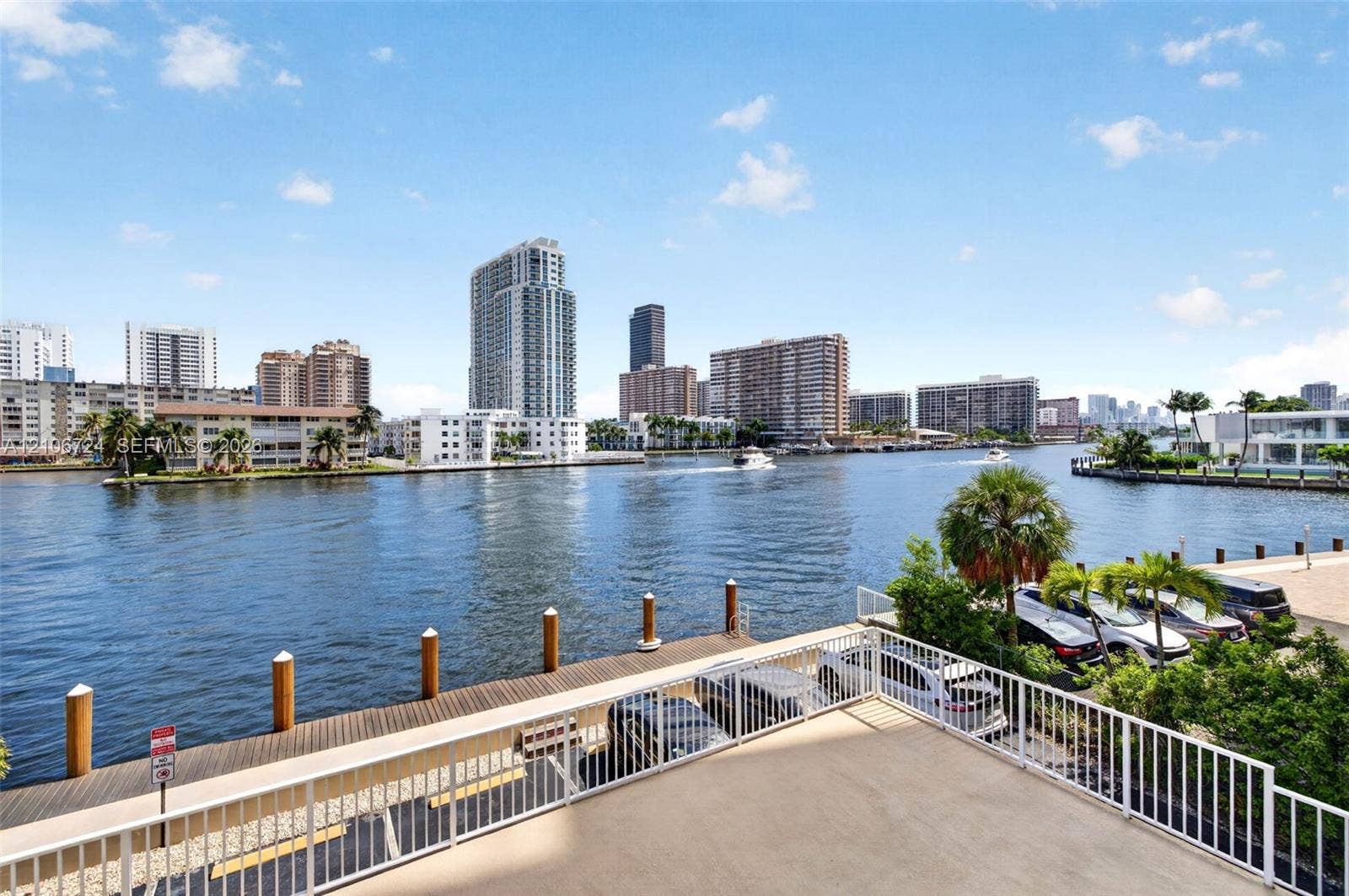 111 Golden Isles Drive, #G12, Hallandale Beach, FL 33009 Concrete balcony with white metal railings, overlooking a waterway, wooden dock, and urban skyline