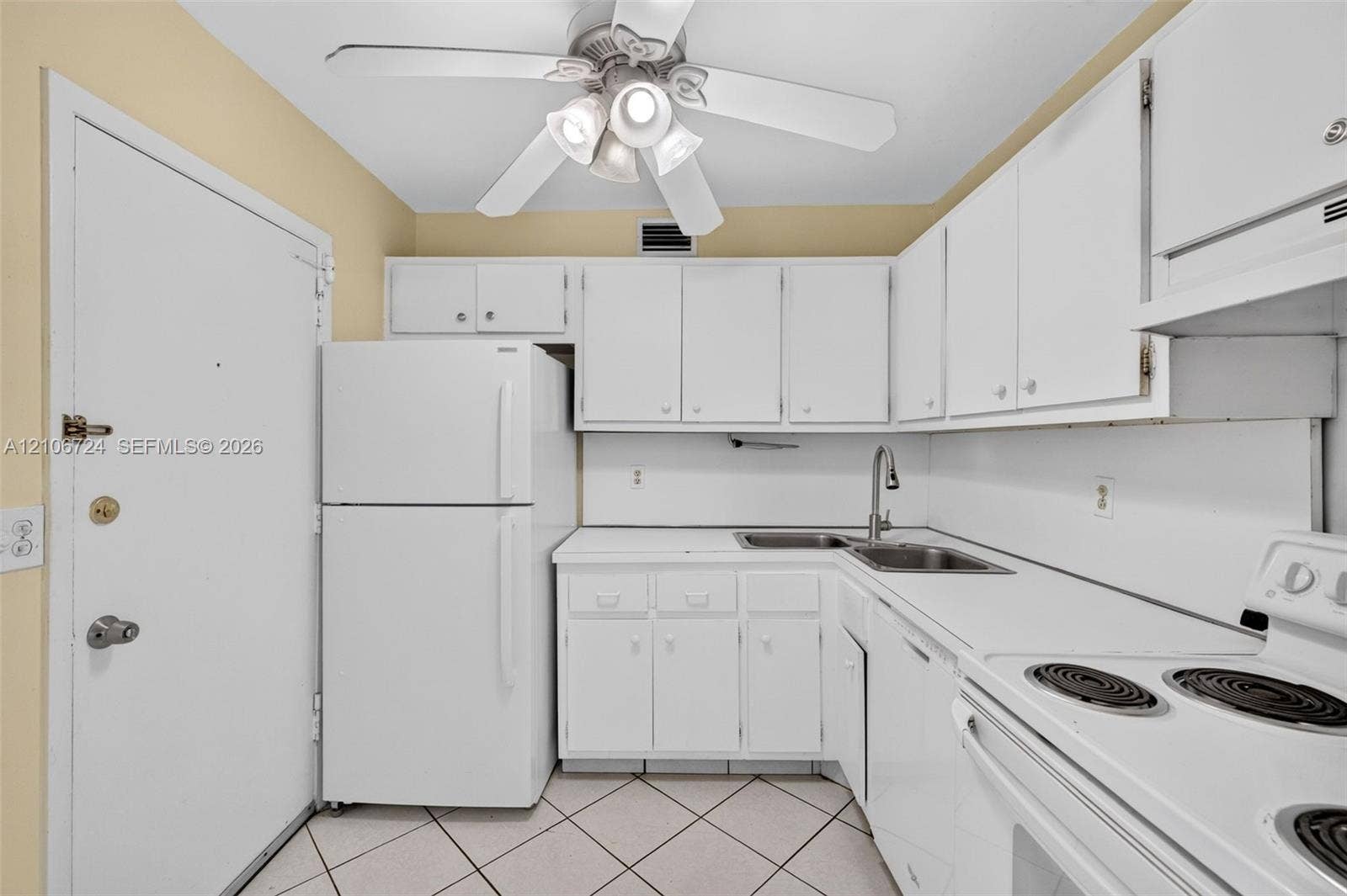 111 Golden Isles Drive, #G12, Hallandale Beach, FL 33009 Kitchen featuring white cabinetry, solid-surface countertops, dual-basin stainless steel sink with high-arc faucet, white appliances, and a ceiling-mounted light fixture with fan