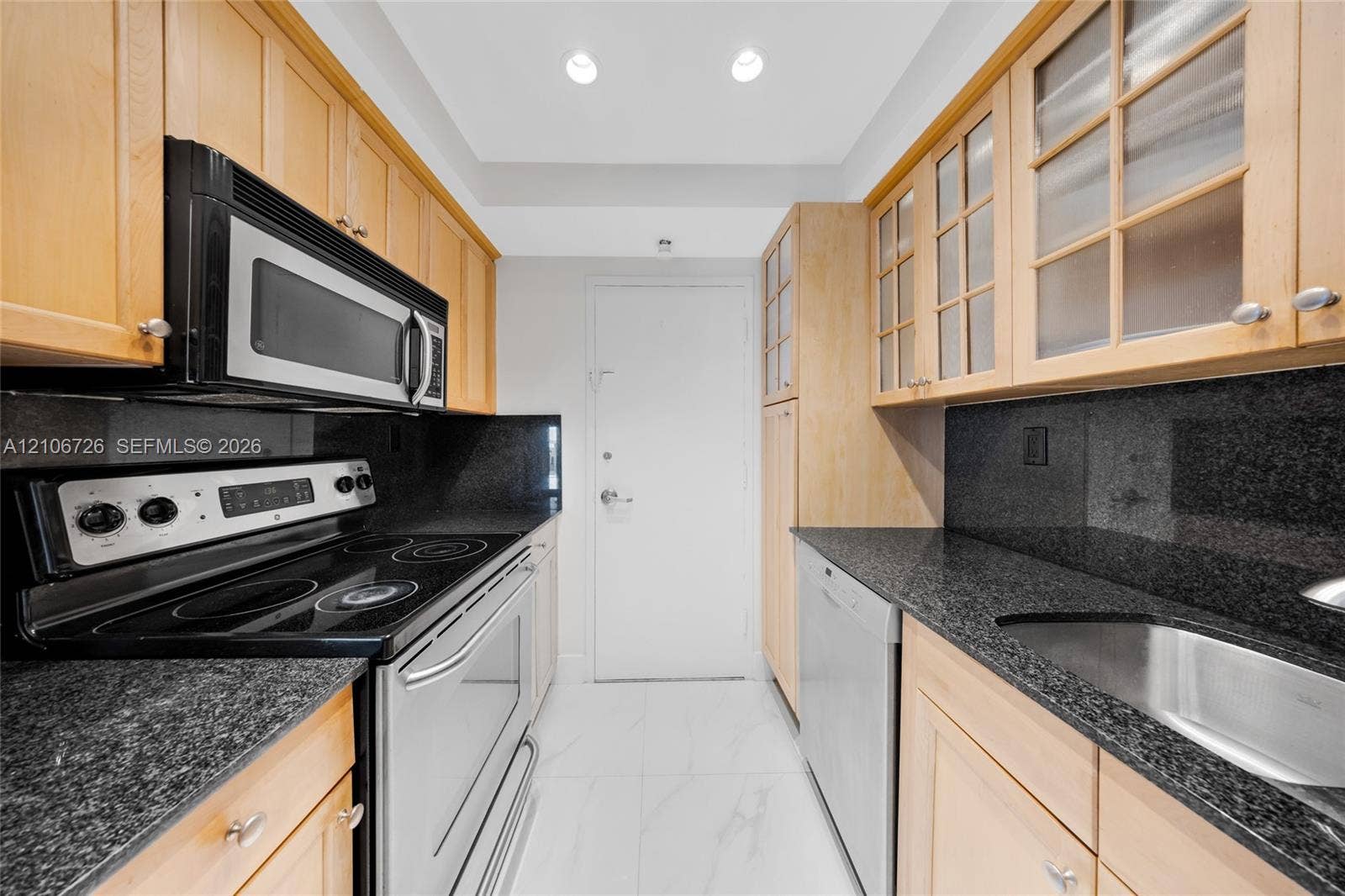 1200 West Avenue, #606, Miami Beach, Miami Beach, FL 33139 Galley kitchen featuring light wood cabinetry with glass-panel upper doors, dark stone countertops, stainless steel appliances, and large-format tile flooring