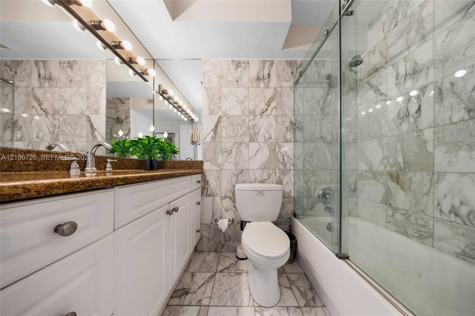 1200 West Avenue, #606, Miami Beach, Miami Beach, FL 33139 Full-width wall mirror with integrated multi-bulb lighting, white cabinetry with metallic hardware, stone-finish countertop, and full-height stone tile surround in the tub-shower combination with a sliding glass door