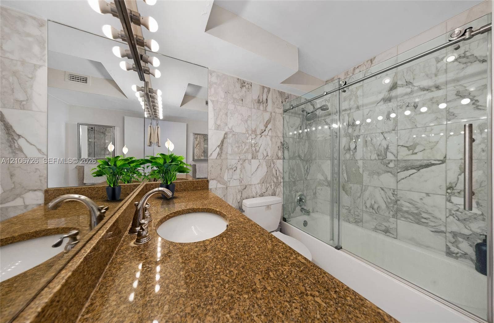 1200 West Avenue, #606, Miami Beach, Miami Beach, FL 33139 Bathroom featuring an expansive vanity with a dual-toned stone countertop, dual undermount sinks, a full-width mirror with integrated lighting, and a bathtub with a stone-tile surround and a glass sliding enclosure