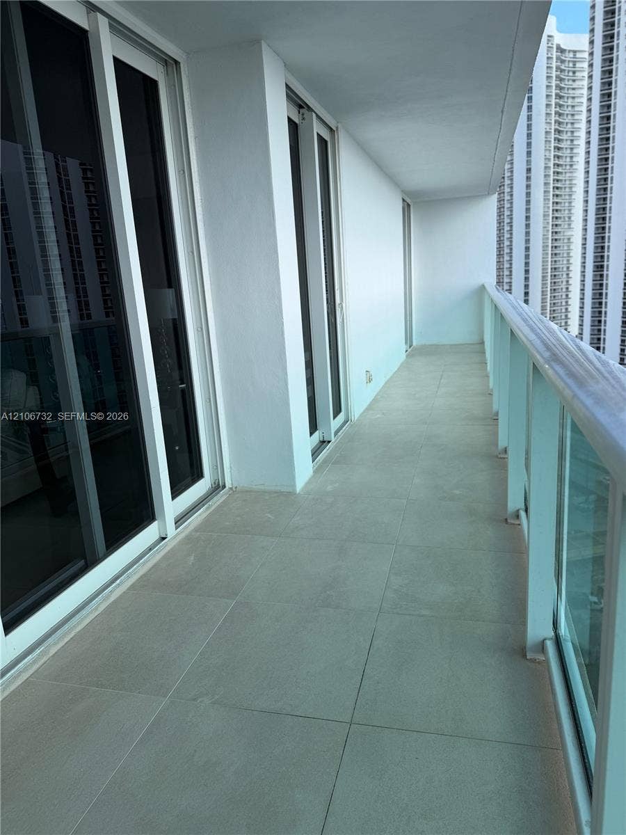 Covered balcony featuring large-format ceramic tile flooring, sliding glass door entry, and glass panel railing with a metal handrail