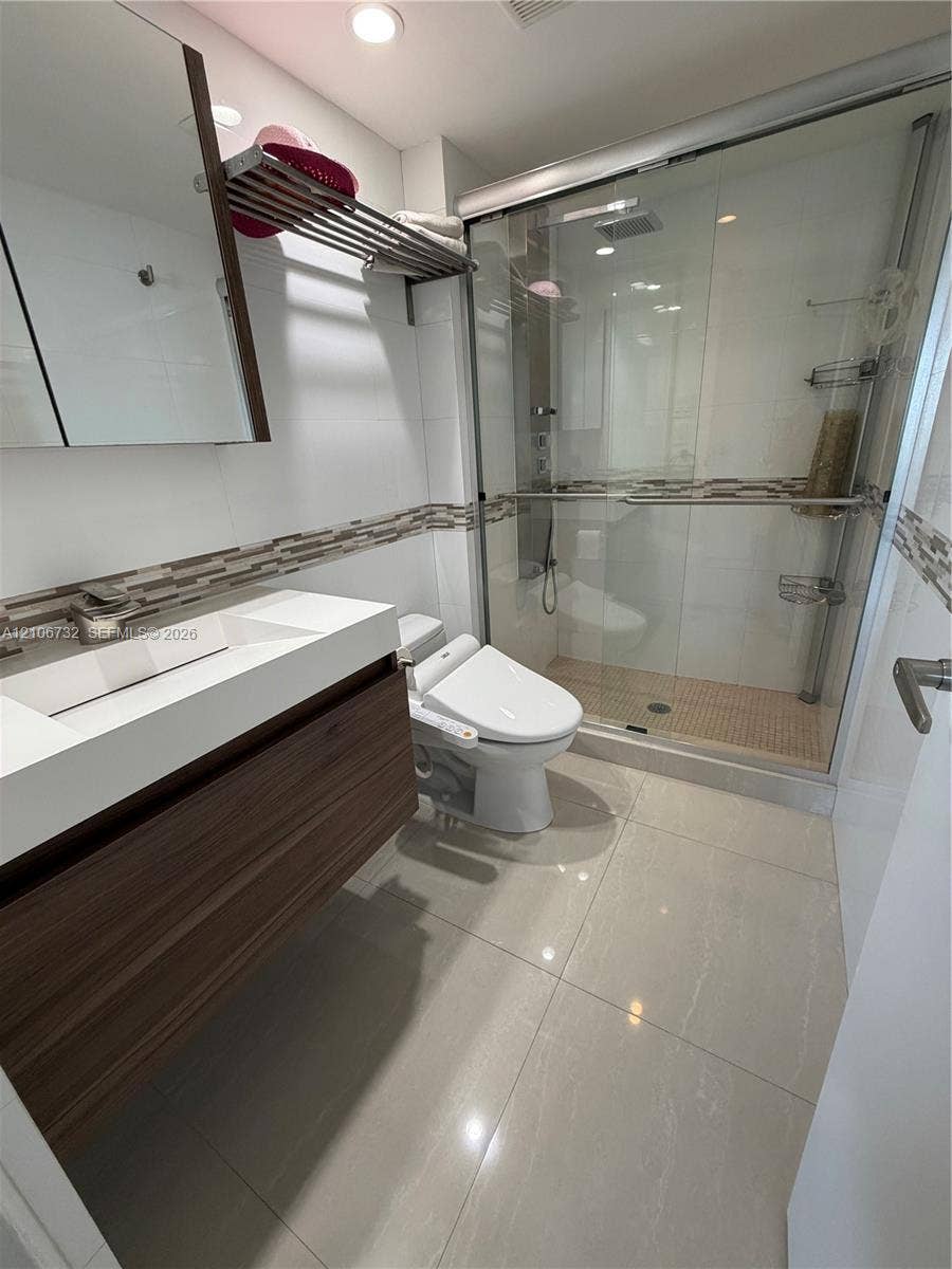 Modern bathroom featuring a wood-finish floating vanity with a white integrated sink, walk-in shower with a glass enclosure, decorative mosaic tile wall accent, and polished large-format floor tiling