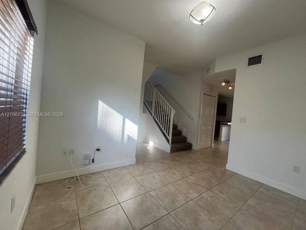 Interior space featuring large-format tile flooring, a central staircase with white railing, ceiling-mounted light fixtures, and windows with horizontal blinds