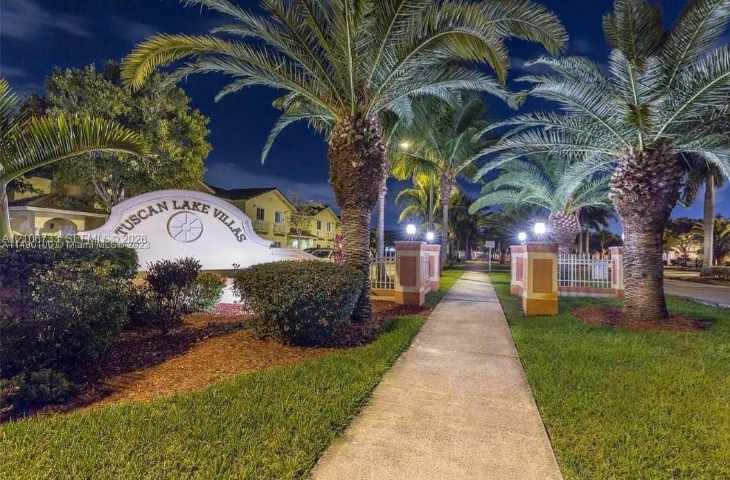 Community signage featuring custom masonry, concrete walkways, professional landscaping, mature palm trees, and decorative post-mounted lighting