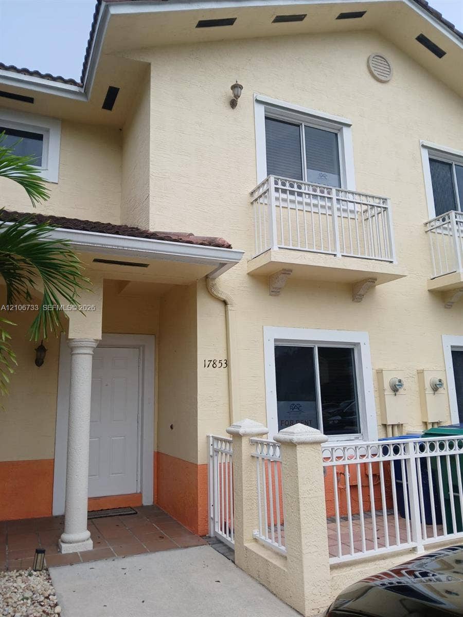 Two-story exterior featuring stucco siding, upper-level balconies with decorative railings, a covered entryway with a column, and tiled flooring