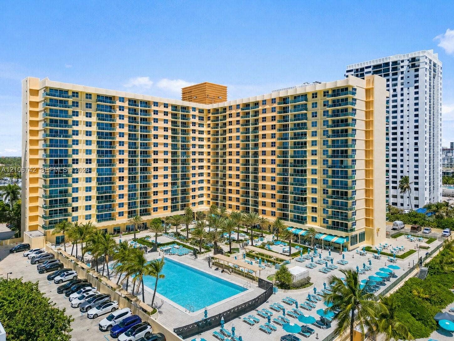 High-rise building featuring uniform balconies with glass railings, outdoor swimming pool with paved terrace, and established palm trees