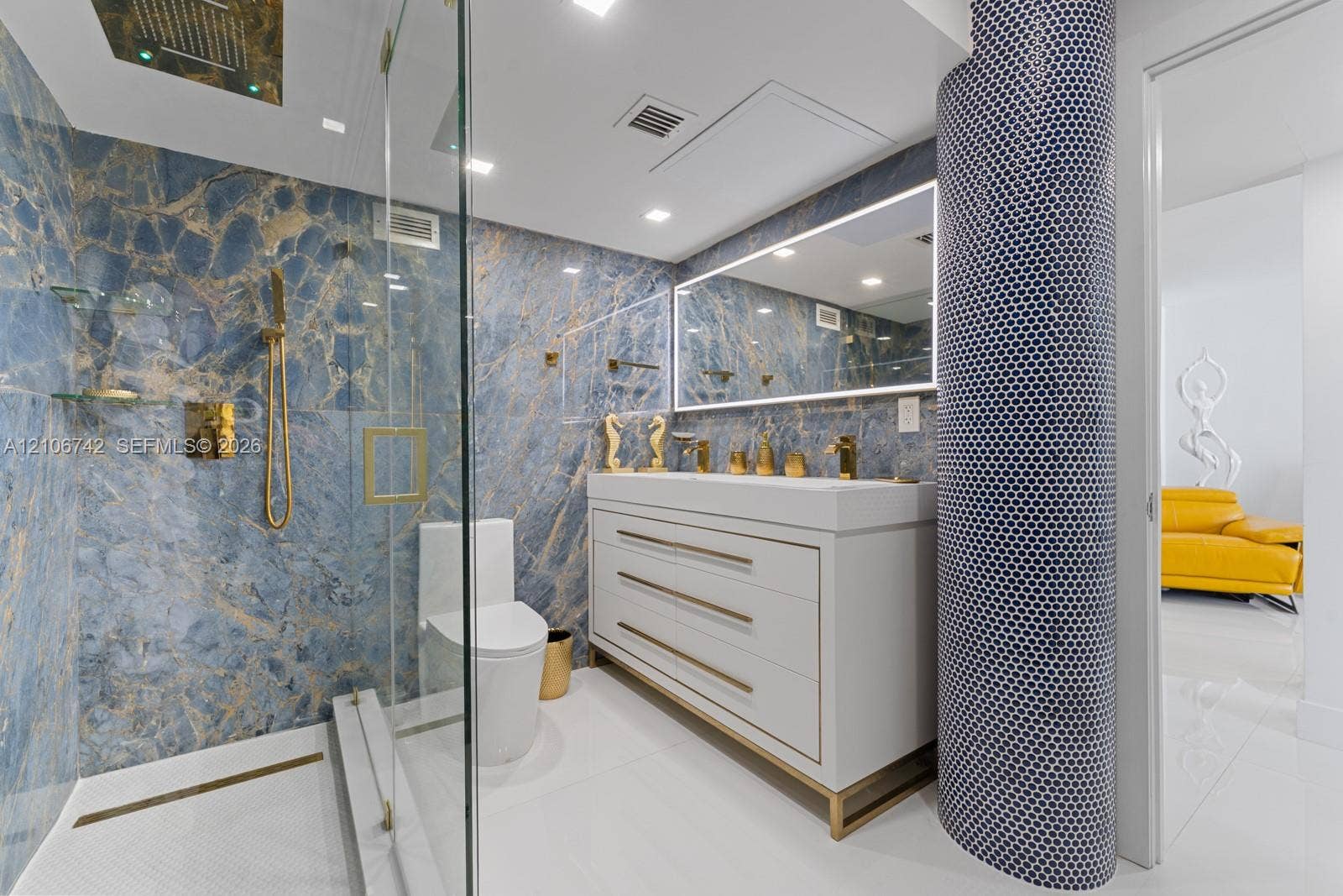 Bathroom featuring blue veined stone wall cladding, a vanity with metallic hardware, a frameless glass-enclosed shower with a rainfall showerhead, and a curved architectural column finished in dark penny-round tile