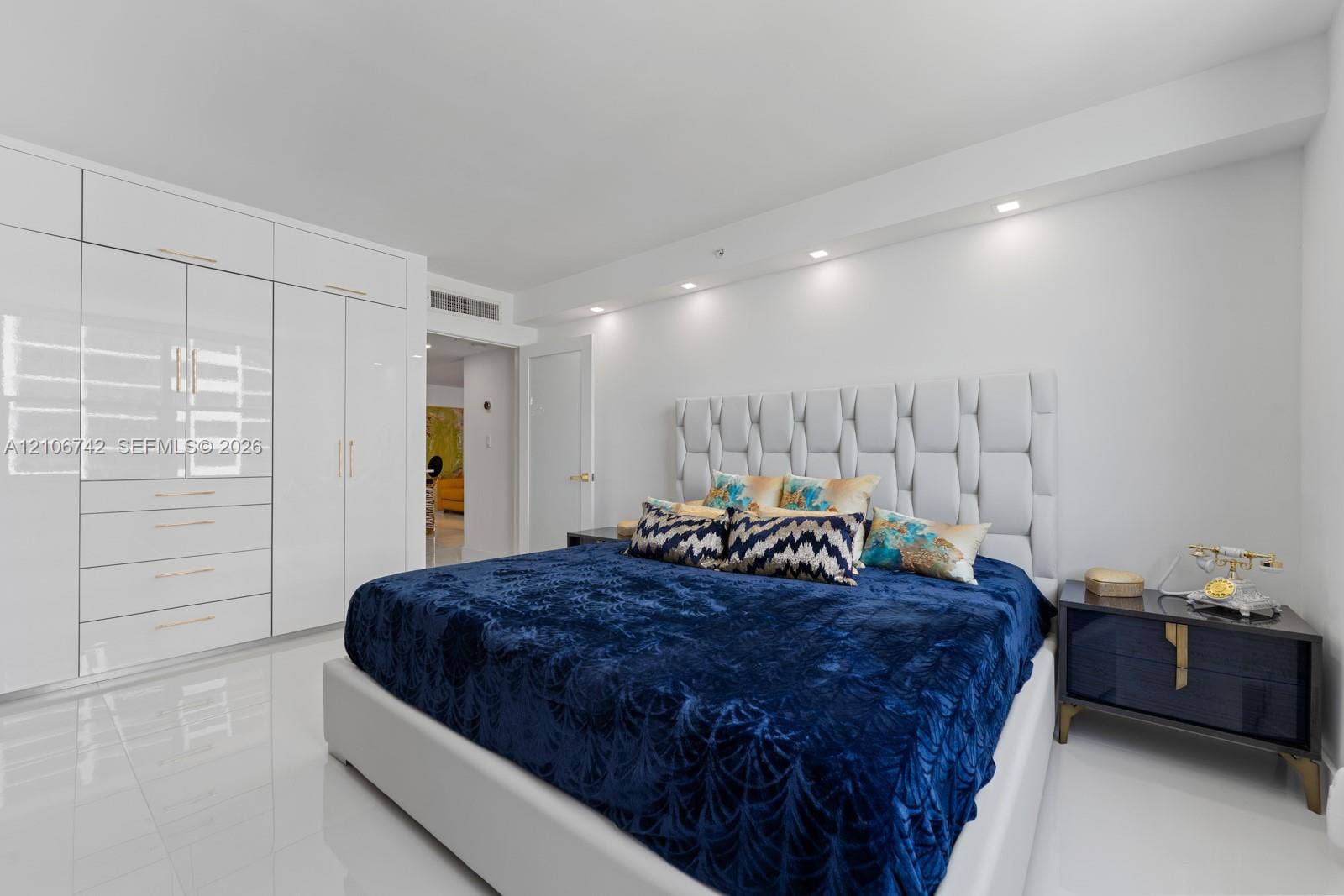 Glossy white floor-to-ceiling built-in cabinetry, polished tile flooring, recessed ceiling lighting, and a custom upholstered wall panel headboard