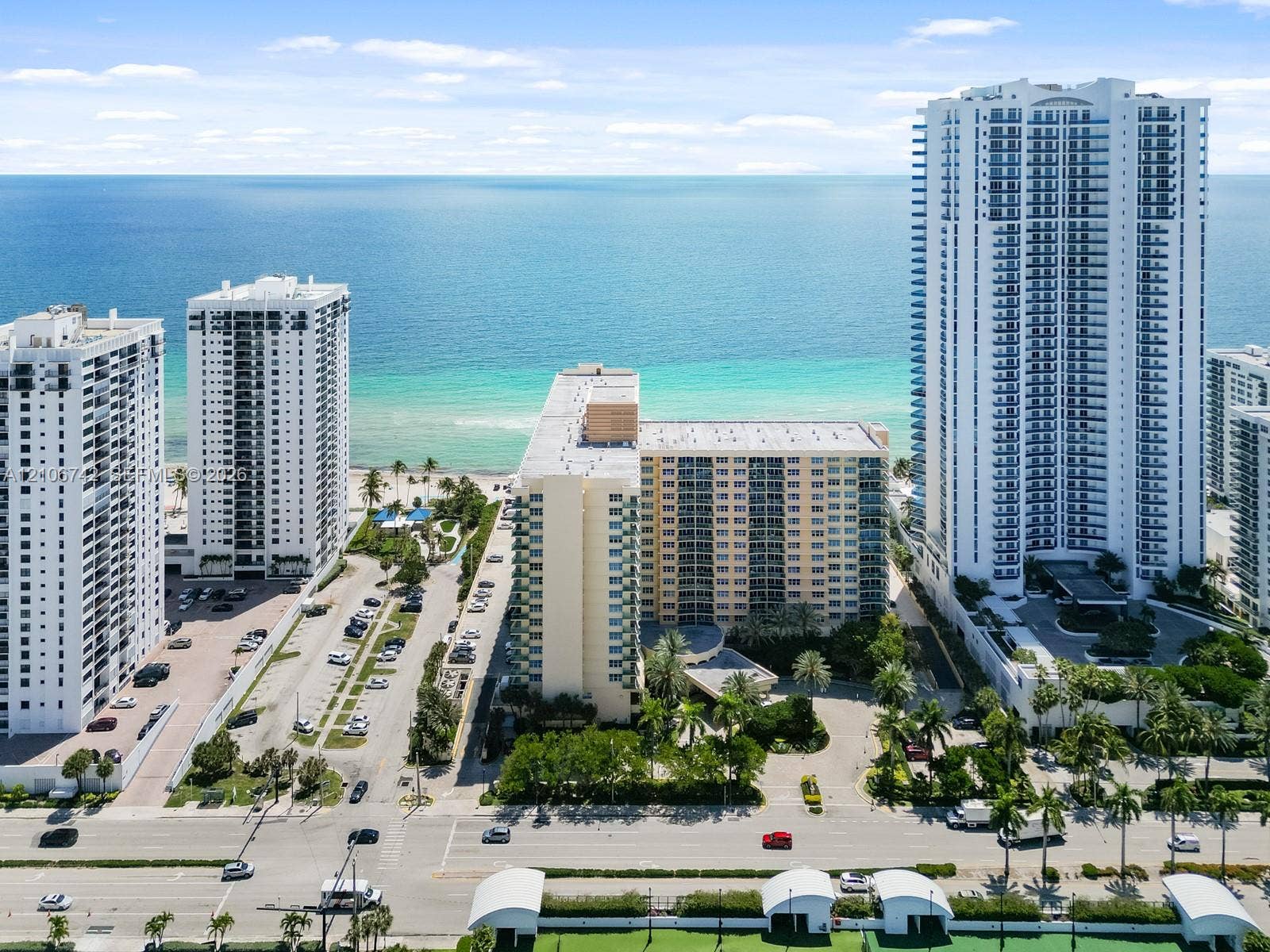 Aerial perspective of high-rise structures situated along a coastline, featuring multi-story architectural facades, paved surface parking, and manicured tropical landscaping