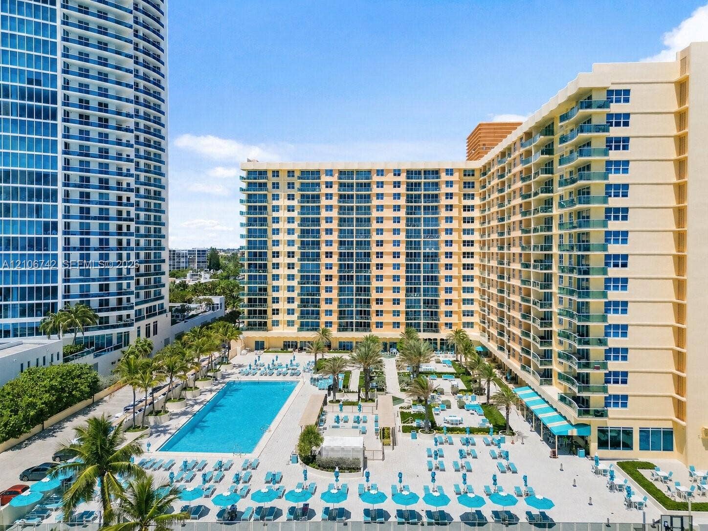 High-rise residential exterior featuring expansive glass balconies, a large rectangular swimming pool, integrated palm tree landscaping, and paved sundeck areas