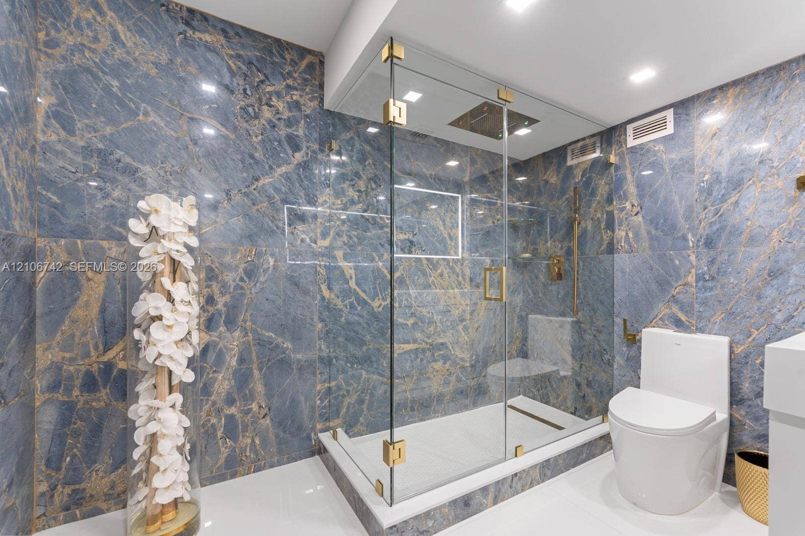 Full-height blue and gold-veined stone wall tiling, glass-enclosed shower with brass hardware, ceiling-mounted rainfall showerhead, and integrated wall niche