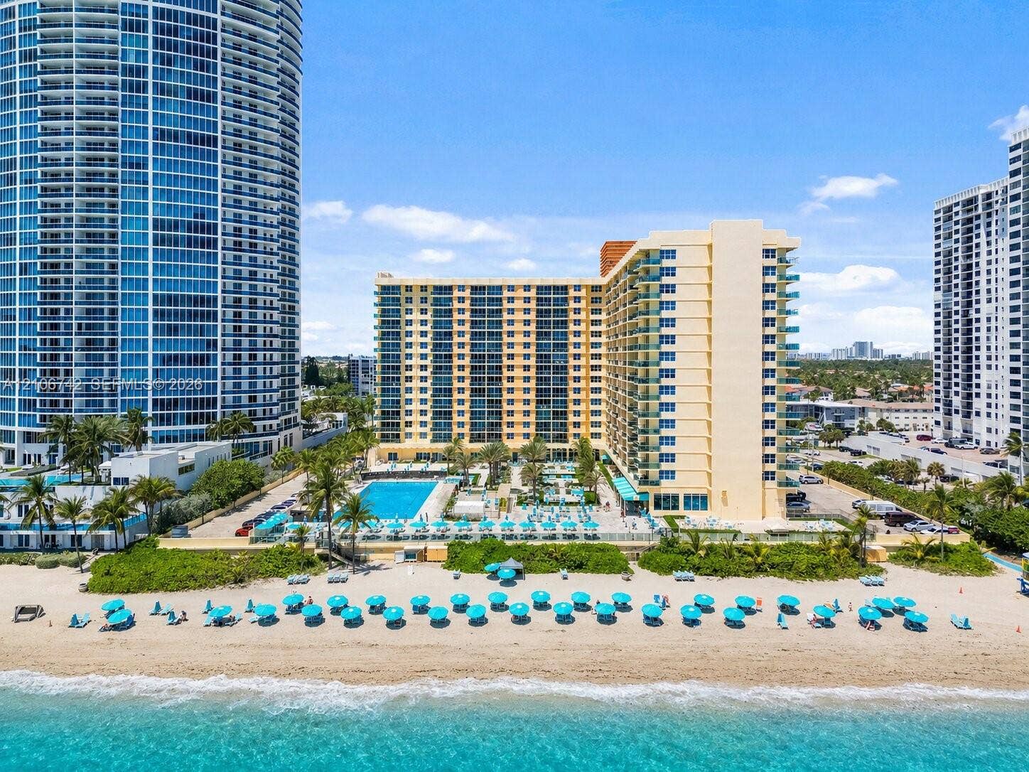 High-rise residential building featuring direct oceanfront access, exterior balconies, an outdoor swimming pool, and landscaped perimeter grounds