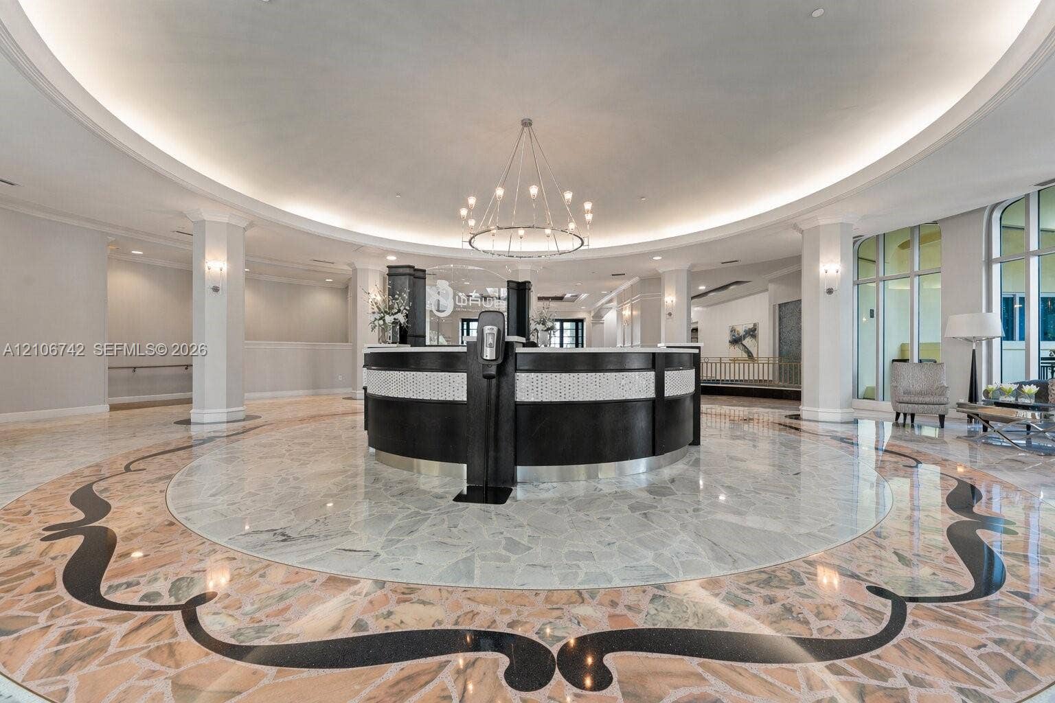 Grand foyer featuring decorative stone flooring with intricate inlays, a curved built-in reception desk, a central multi-arm chandelier, recessed ceiling cove lighting, and structural columns