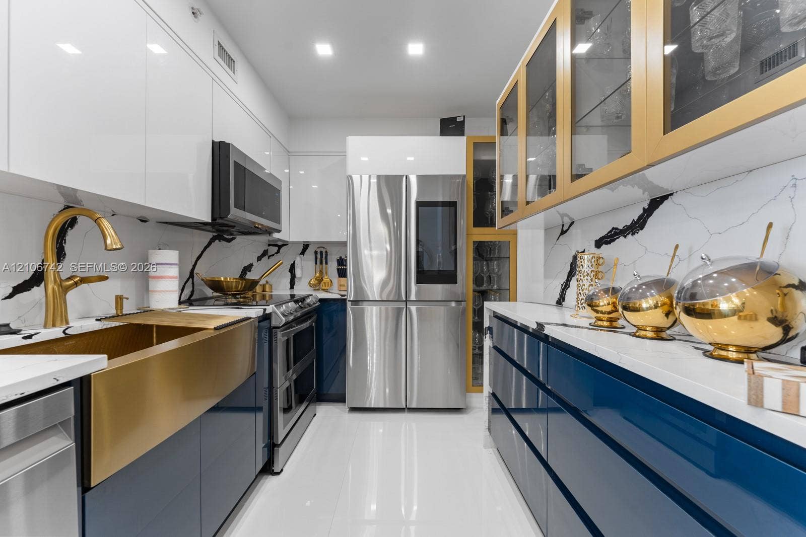 Galley kitchen featuring high-gloss blue and white cabinetry, stone-surfaced countertops with decorative veining, brass-finished farmhouse sink, gold-tone accent cabinetry, and stainless steel appliances