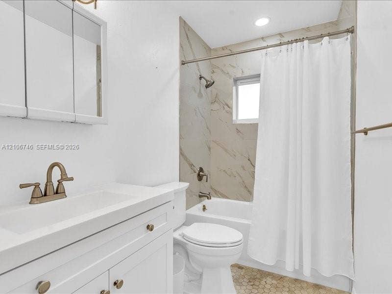 Bathroom featuring a white vanity cabinet, rectangular undermount sink with a bronze-finish faucet, recessed lighting, tub-shower combination with floor-to-ceiling stone-pattern tile surround, and hexagonal tile flooring