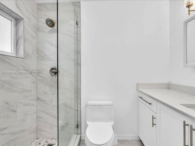 Walk-in shower with frameless glass enclosure, floor-to-ceiling stone-tile surround, vanity with stone-composite countertop, and wall-mounted light fixture