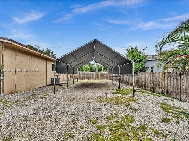 Metal-framed carport with a pitched canopy, positioned over a gravel surface and enclosed by a wooden privacy fence