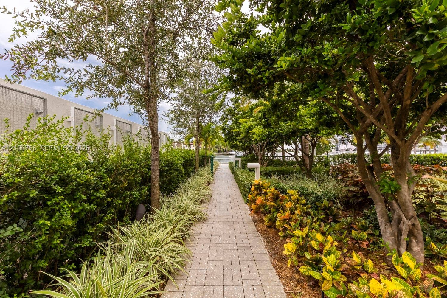 Paver walkway lined with mature trees, manicured hedges, and ornamental tropical landscaping