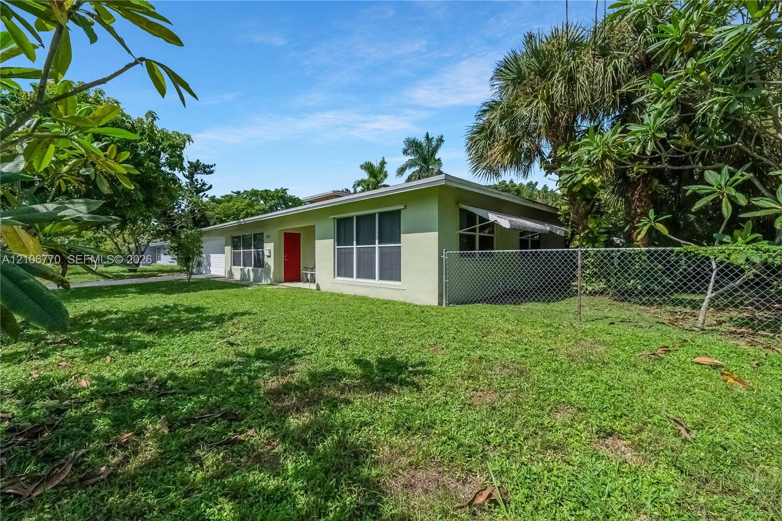 5701 SW 1st Street, Plantation, FL 33317 Single-story residence featuring light-colored stucco siding, a bold red entry door, large grid-pattern windows, and a chain-link fenced perimeter