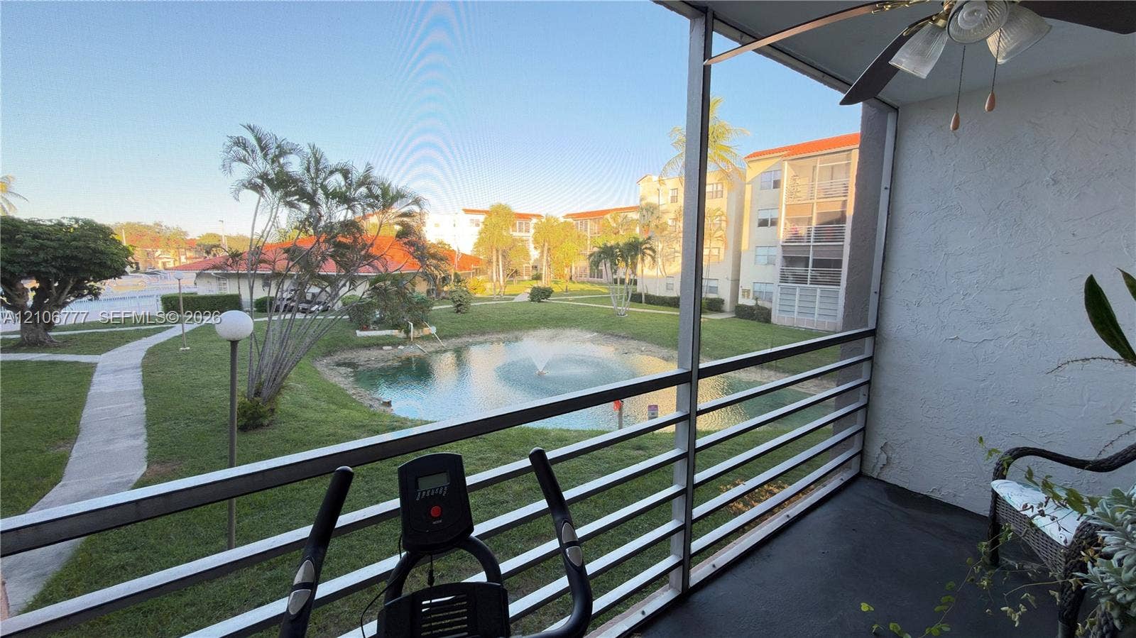 1810 SW 81st Avenue, #2200, North Lauderdale, North Lauderdale, FL 33068 Screened-in balcony with horizontal metal railing, ceiling-mounted fan, and exterior lighting fixture, overlooking a landscaped pond with a fountain