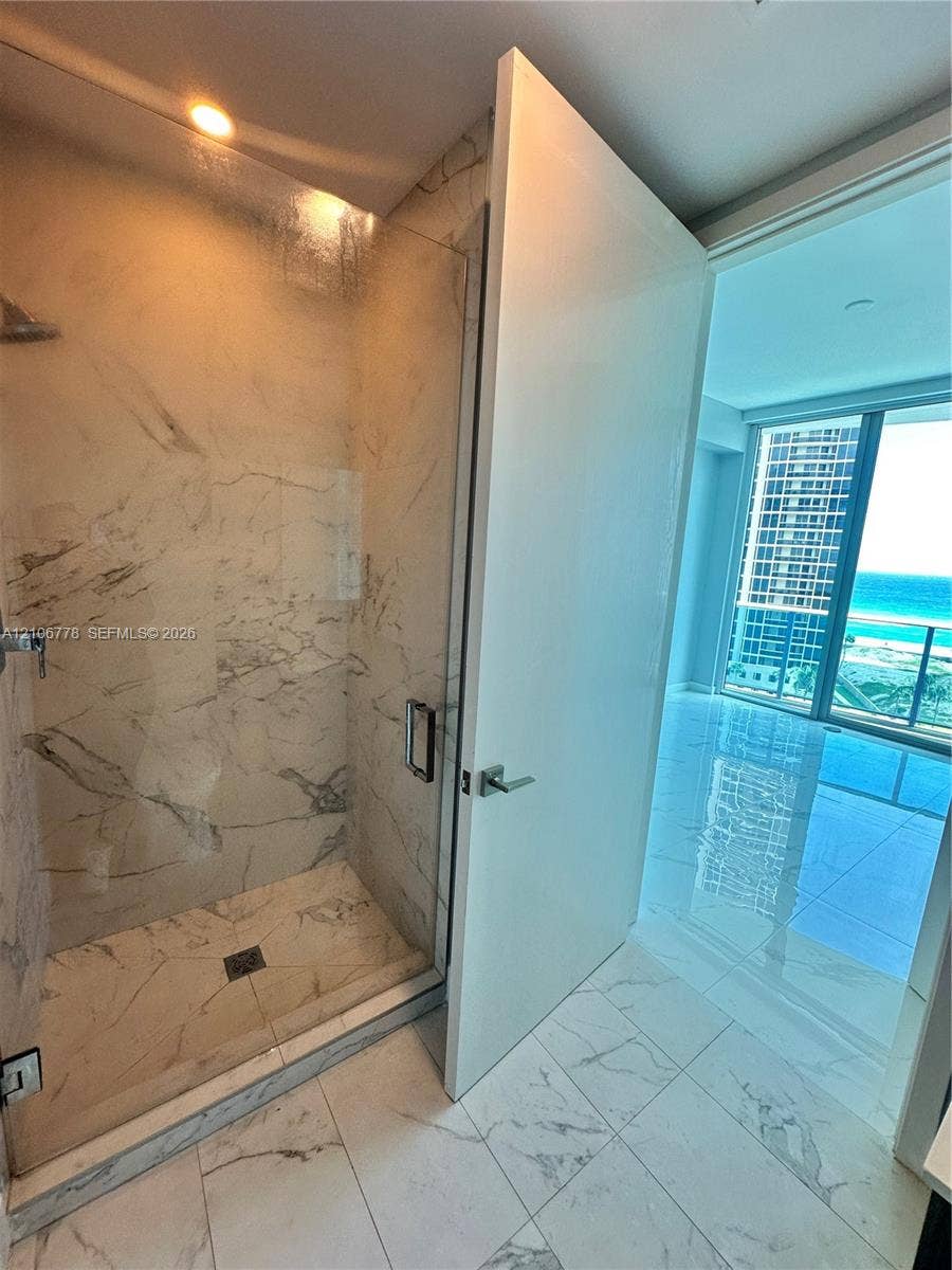 17550 Collins Avenue, #803, Sunny Isles Beach, Sunny Isles Beach, FL 33160 Walk-in shower featuring glass enclosure, ceiling-mounted fixture, and veined stone-finish wall tile, complemented by large-format polished flooring throughout the suite and floor-to-ceiling glass