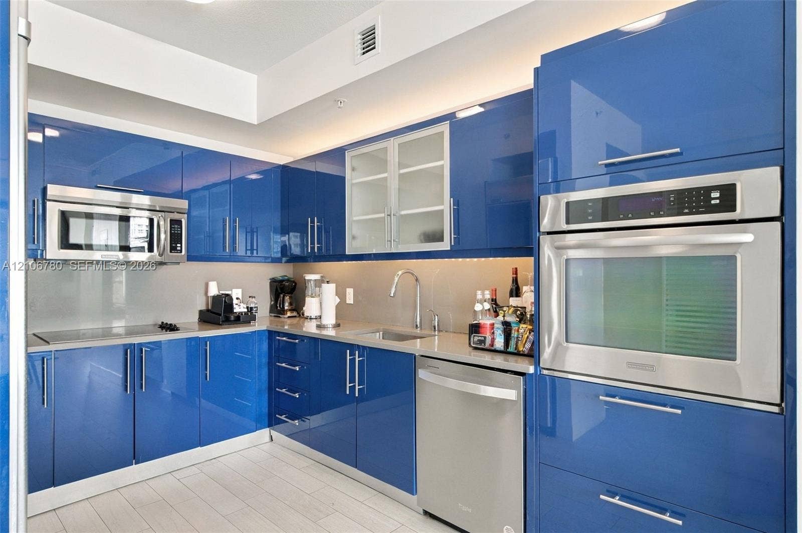 Glossy blue cabinetry, high-gloss flat-panel doors, stainless steel wall-mounted oven and microwave, neutral stone-finish countertops, and stainless steel undermount sink