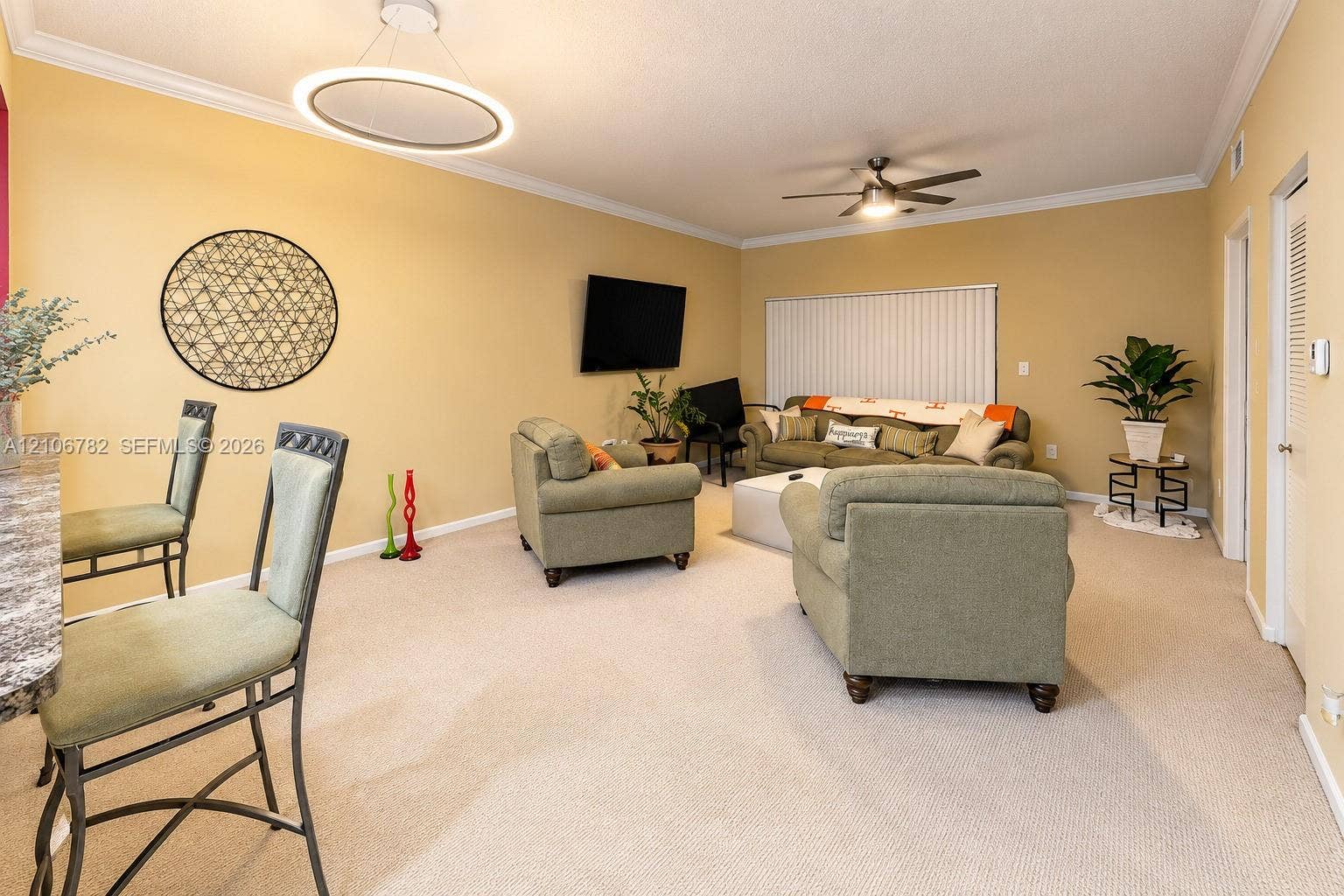 Living area featuring neutral carpeting, a modern circular LED pendant light, crown molding, a ceiling fan with integrated lighting, and vertical window blinds