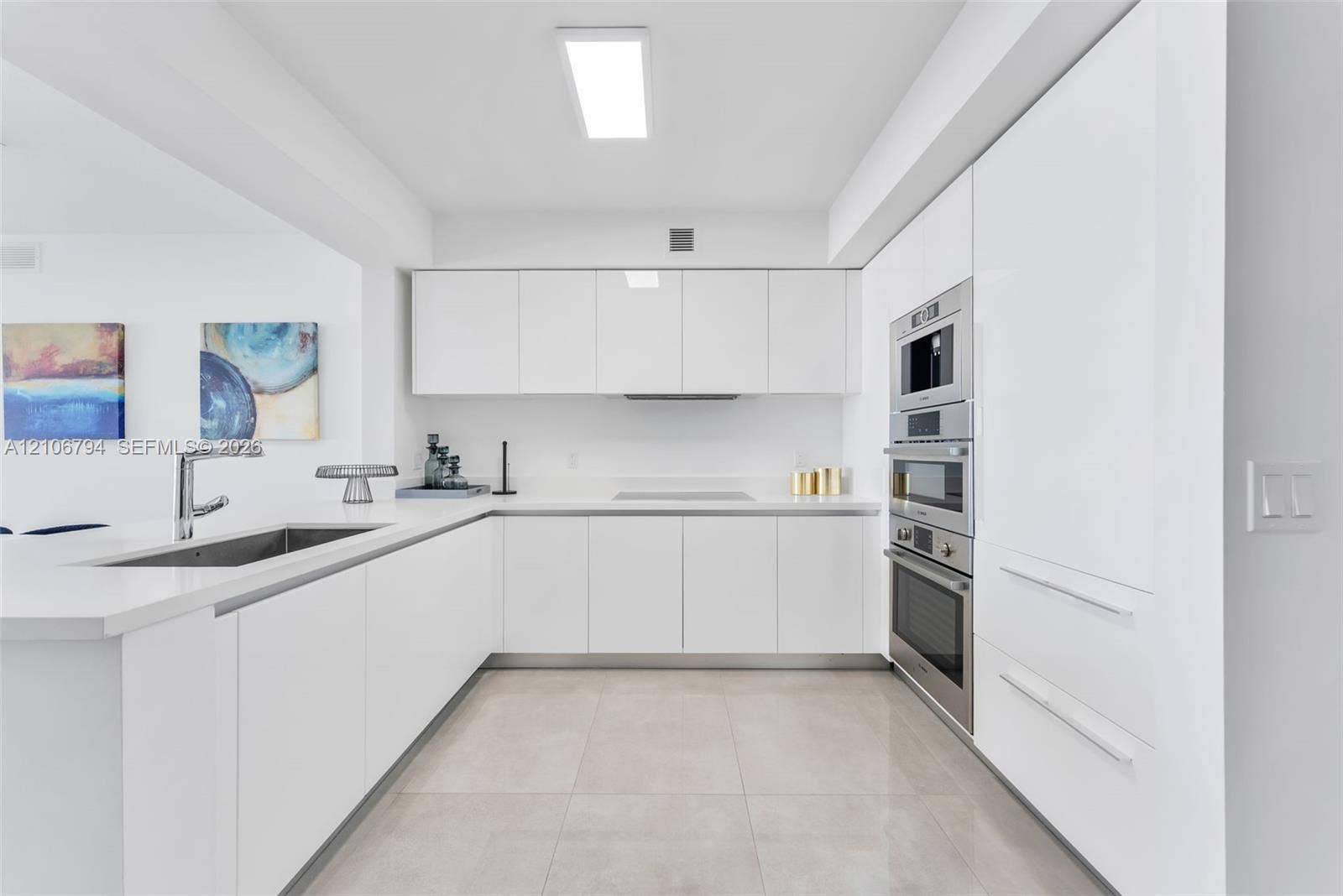 Modern kitchen featuring white flat-panel cabinetry, light-toned tile flooring, integrated stainless steel appliances, and solid-surface countertops with an undermount sink