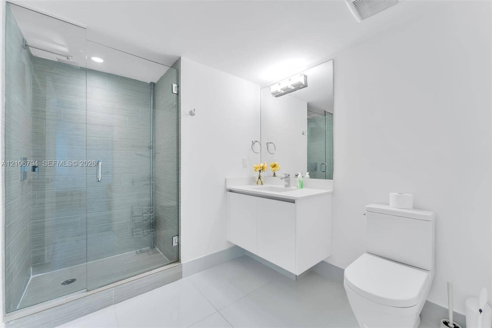 Walk-in shower with horizontal tile surround and glass door, wall-mounted white vanity, solid-surface countertop with integrated sink, and large-format tile flooring