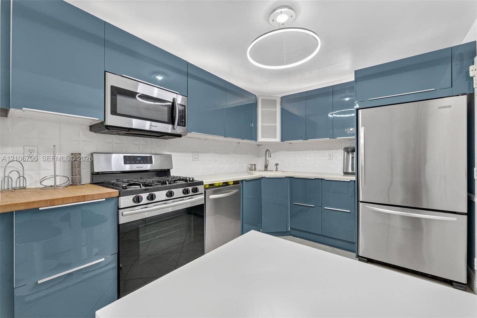 Glossy blue cabinetry featuring integrated hardware, stainless steel appliances, solid surface countertops, textured backsplash tiling, and a circular LED flush-mount light fixture
