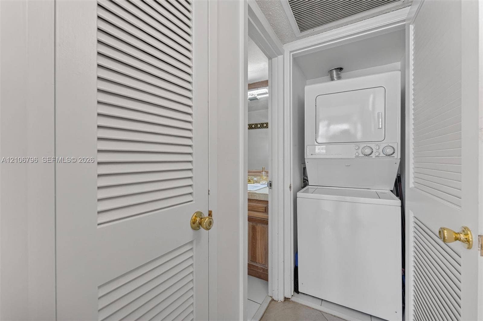 Stacked laundry unit featuring white cabinetry, louvered closet doors with brass-finish hardware, and tiled flooring