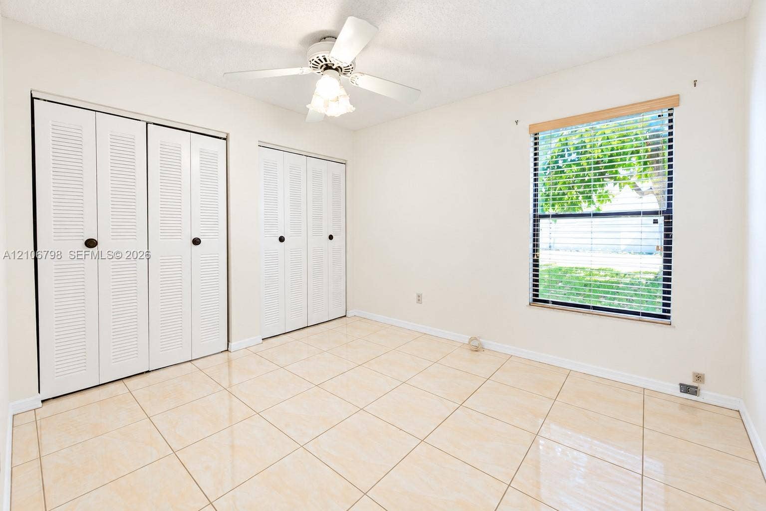14750 SW 156th Street, Miami, FL 33187 Interior room featuring large-format neutral tile flooring, dual louvered-style closets, a white ceiling fan with integrated light fixture, and a single window with wood-finish trim