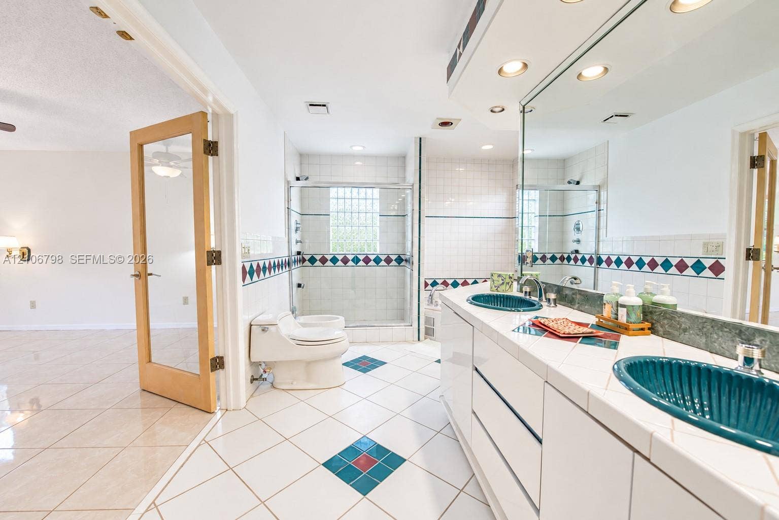 14750 SW 156th Street, Miami, FL 33187 Bathroom featuring a dual-sink vanity with tiled countertops, two ribbed teal vessel sinks, a glass-enclosed shower with glass block window, and white tile flooring with decorative accent insets