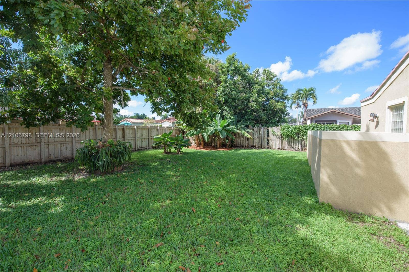 14750 SW 156th Street, Miami, FL 33187 Backyard featuring a manicured lawn, perimeter wood fencing, mature trees, and an exterior masonry wall