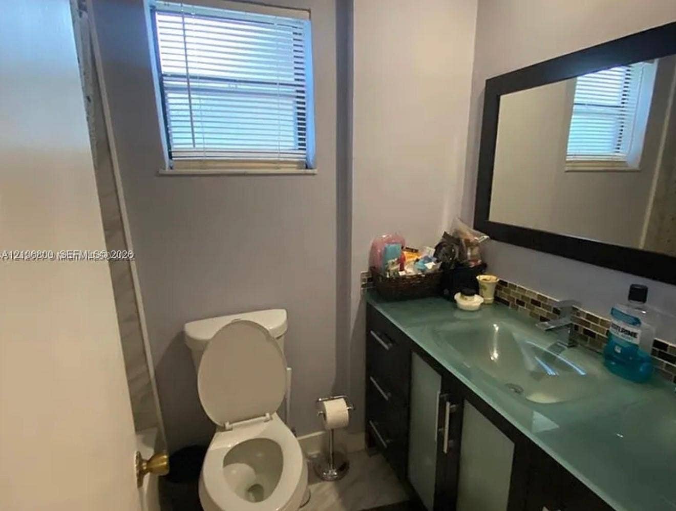 3820 SW 52nd Avenue, Hollywood, Hollywood, FL 33023 Bathroom featuring a dark-finish vanity with glass-panel cabinet doors, an integrated glass countertop with a molded sink, a dark-framed wall mirror, and a window with integrated blinds