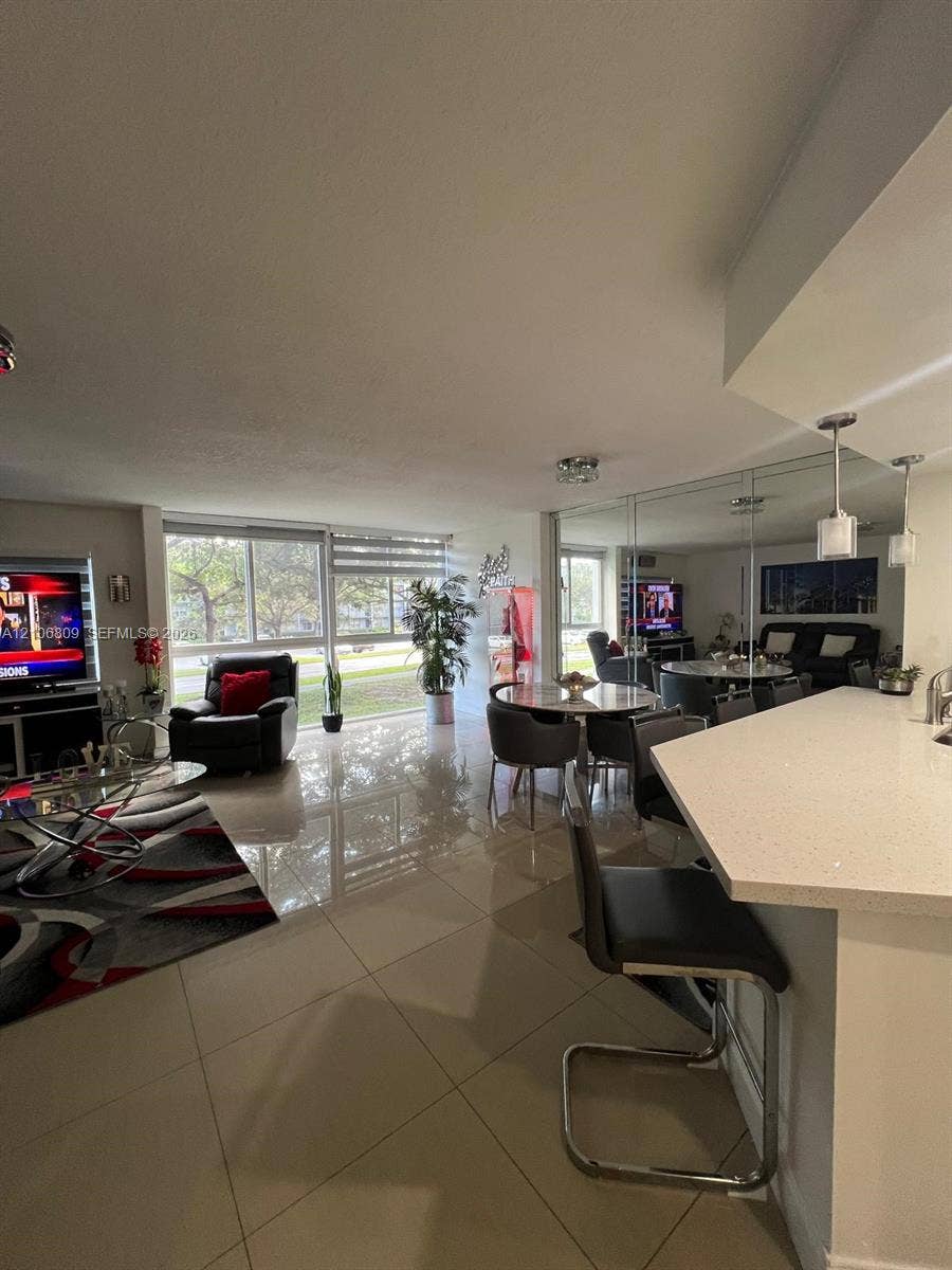 12600 SW 5th Court, #204L, Pembroke Pines, FL 33027 Open-concept living space featuring large-format ceramic tile flooring, a kitchen breakfast bar with stone-surfaced countertops, floor-to-ceiling mirrored wall accents, pendant lighting, and oversized sliding windows