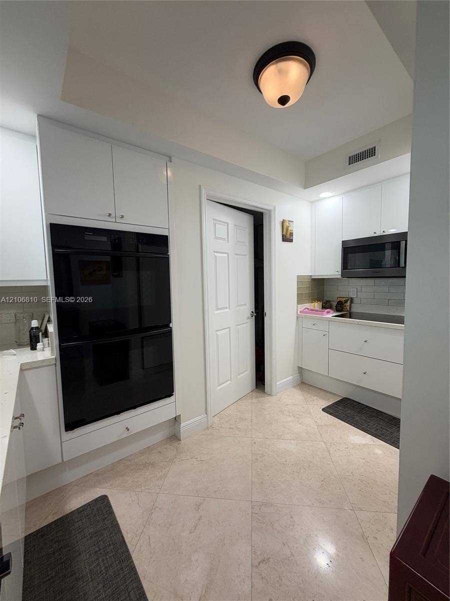 Kitchen featuring white flat-panel cabinetry, a built-in double oven, over-the-range microwave, light-toned stone-finish tile flooring, and a subway tile backsplash