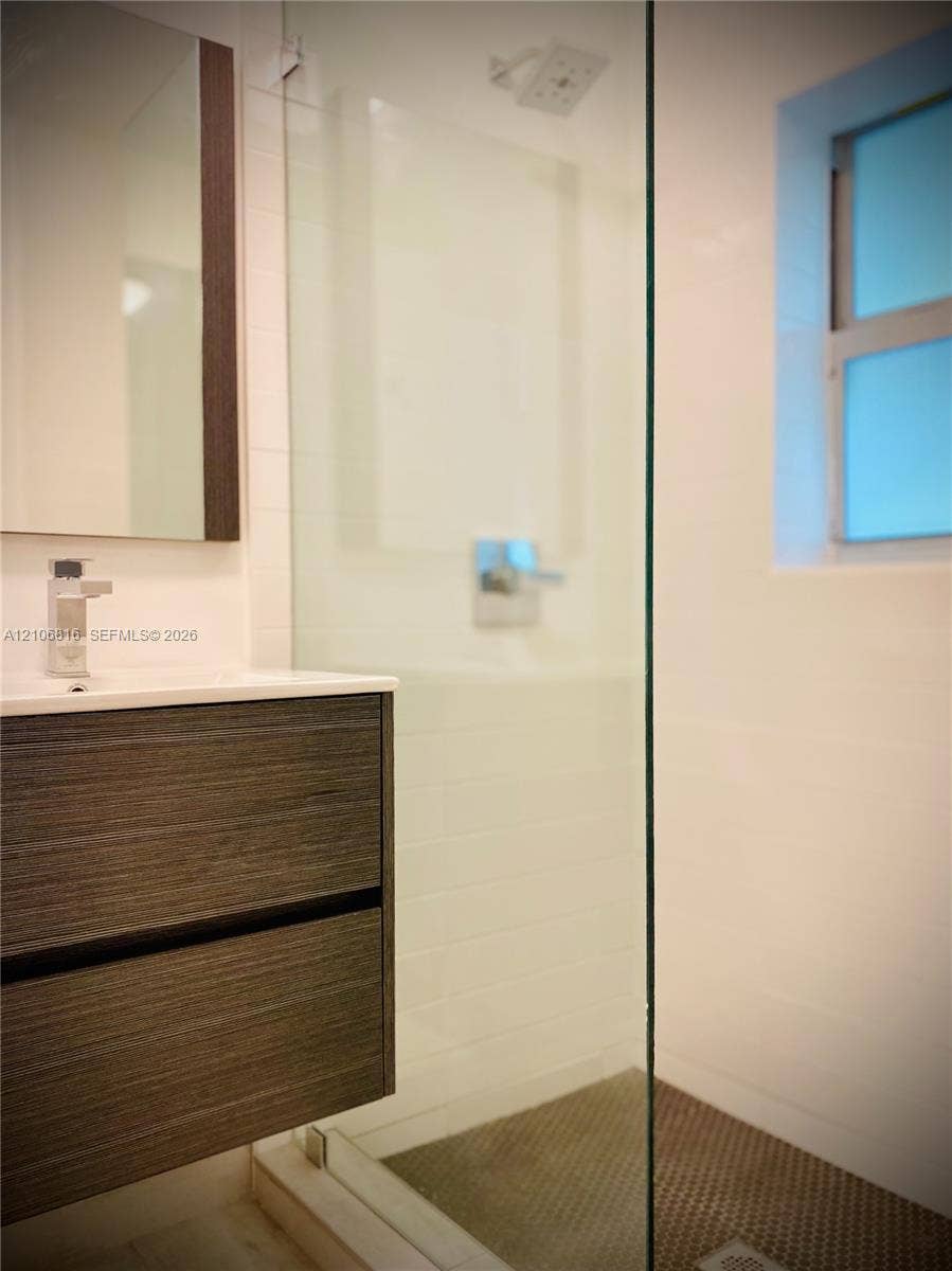 Floating wood-finish vanity with a rectangular sink, wall-mounted mirror, glass-enclosed shower featuring a rainfall showerhead, and mosaic tile flooring
