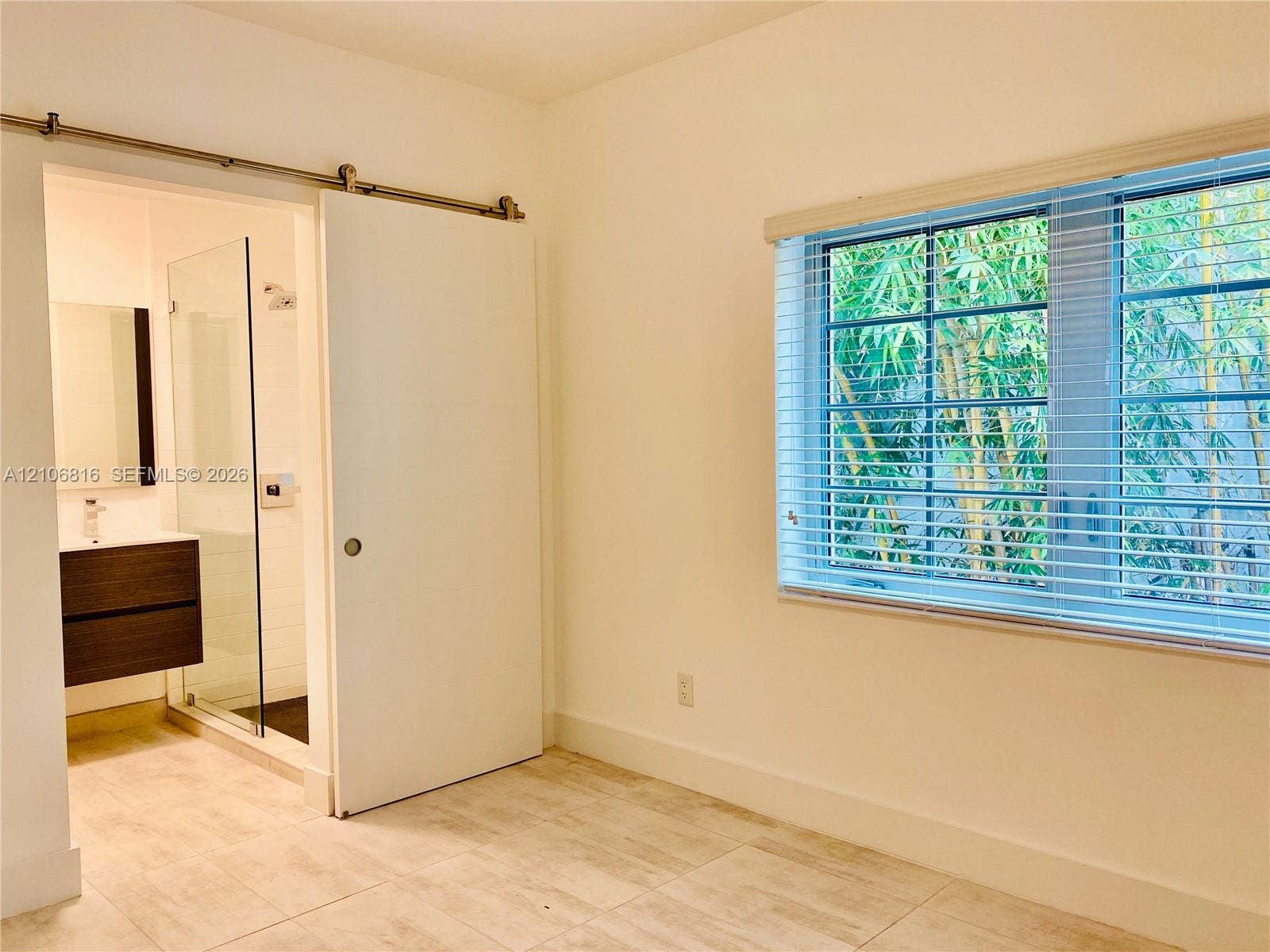 Interior space featuring tile flooring, sliding barn door with metal hardware, multi-pane window with horizontal blinds, and attached bathroom with floating vanity and glass-enclosed shower