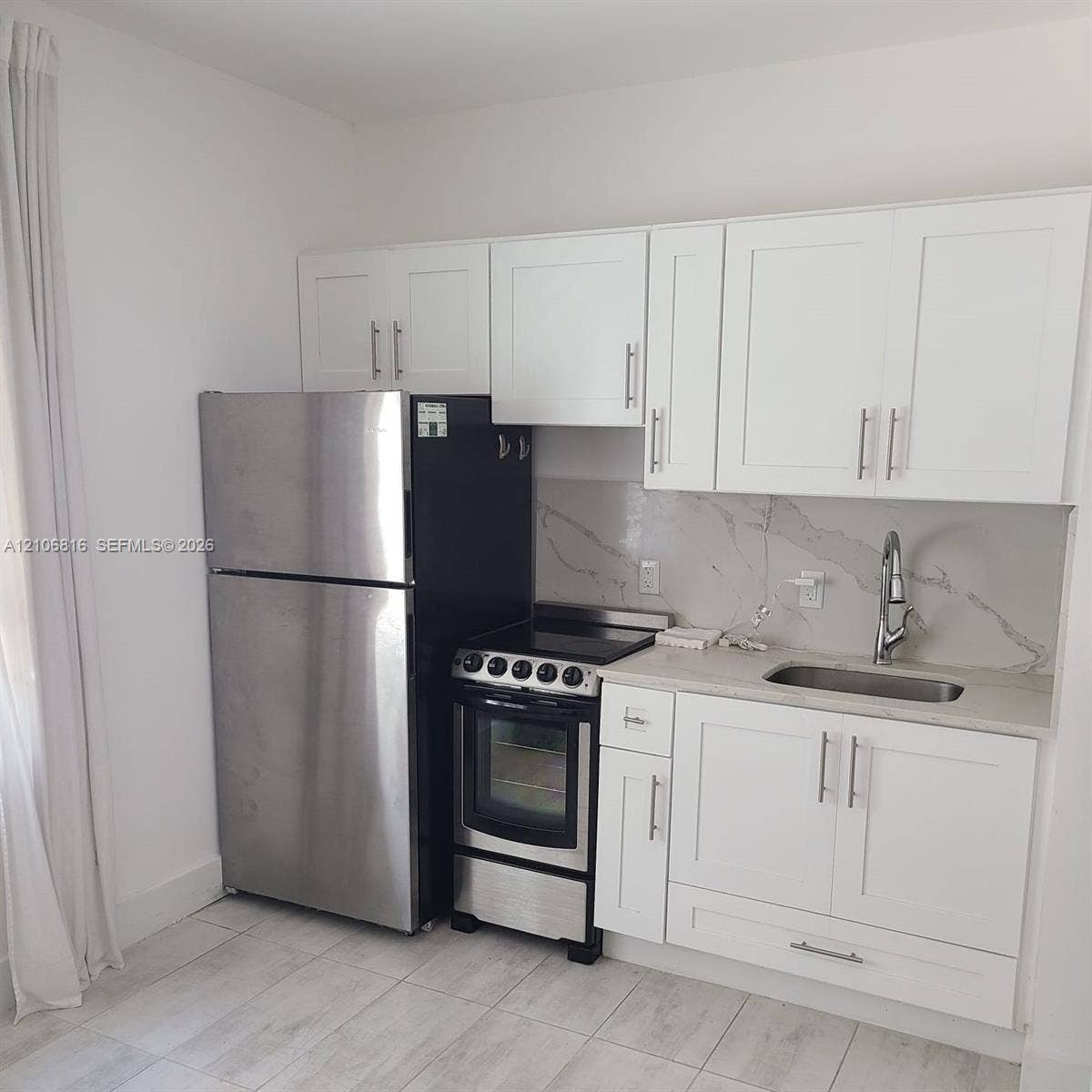 Kitchen area featuring white shaker-style cabinetry, a stone-patterned backsplash, stainless steel refrigerator and range, and light-toned tile flooring