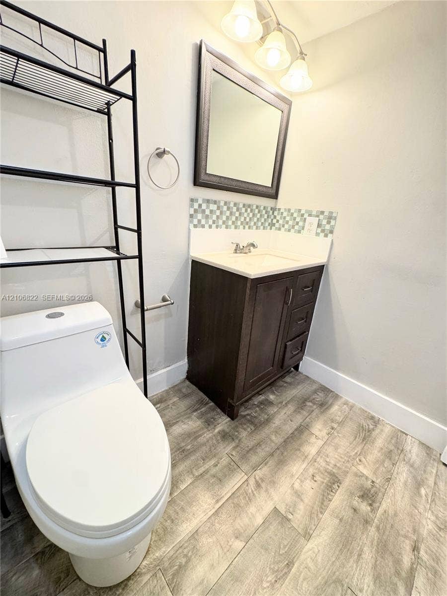 Dark-finish vanity with a rectangular sink, patterned glass mosaic backsplash, wood-finish flooring, and a framed mirror