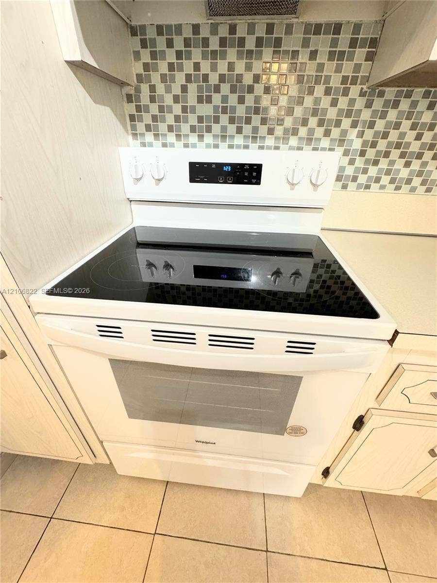 White Whirlpool electric range with a smooth glass cooktop and digital control panel, complemented by a multi-tonal square tile backsplash and tile flooring