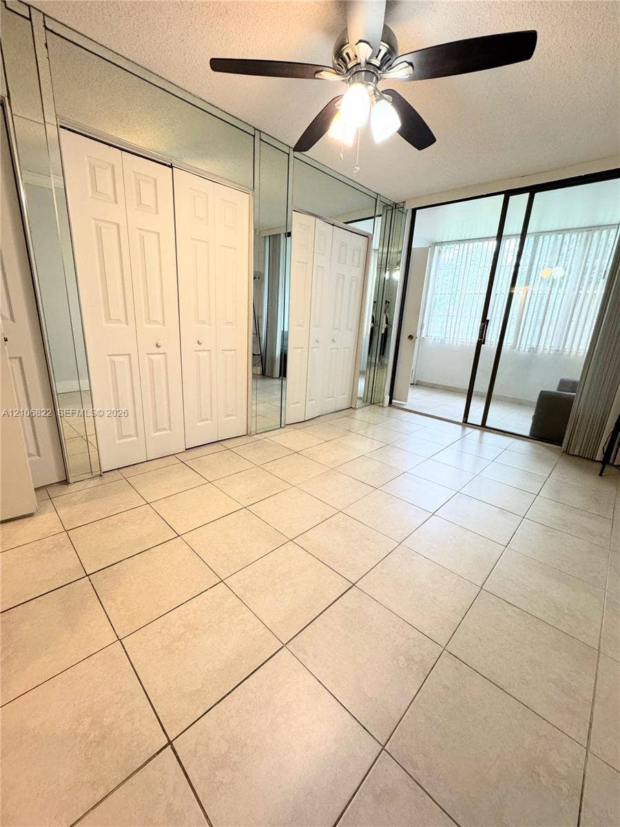 Ceramic tile flooring, floor-to-ceiling mirrored closet doors, and sliding glass patio doors with vertical blinds, finished with a ceiling fan with integrated light fixture
