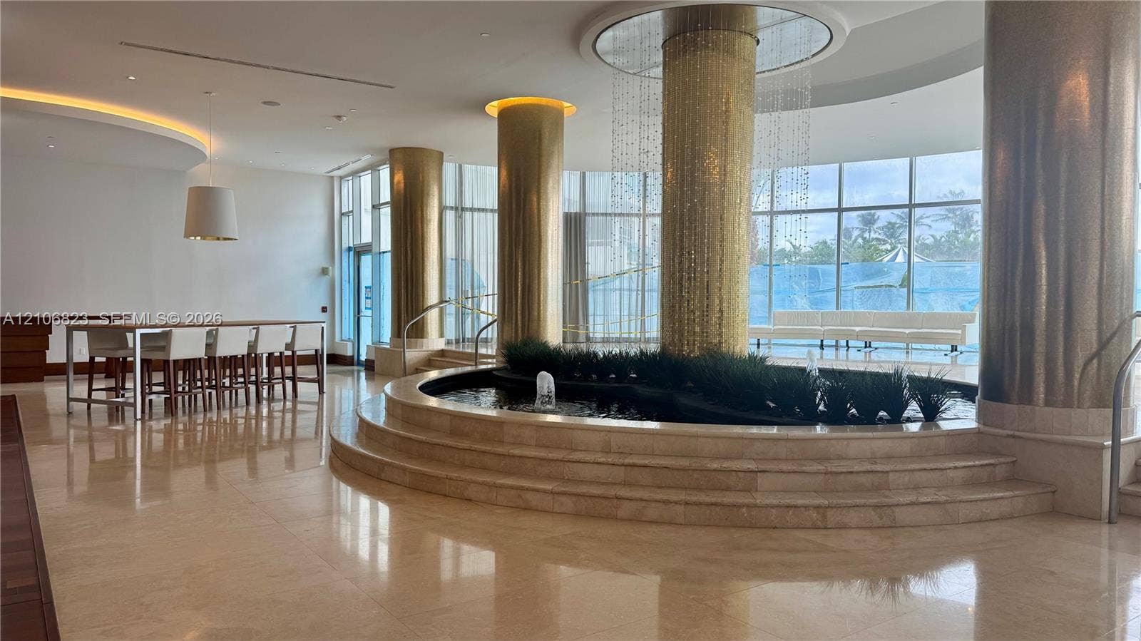 Grand lobby featuring polished stone flooring, circular tiered water feature, gold-tiled support columns, full-height floor-to-ceiling windows, and recessed cove lighting