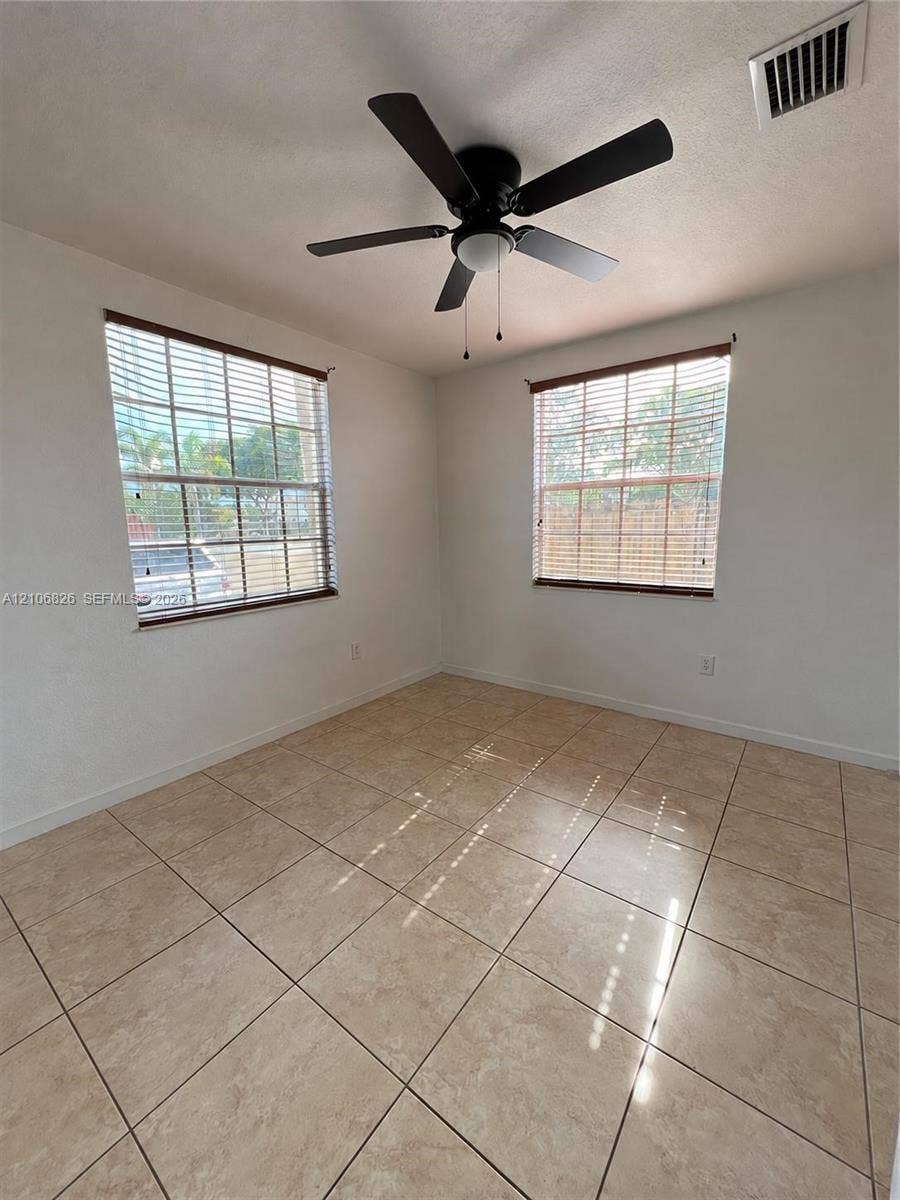 Room featuring neutral tile flooring, dual windows with horizontal blinds, and a central ceiling fan with integrated lighting