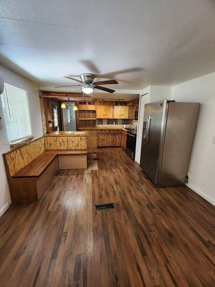 Kitchen featuring wood-finish flooring, custom wood cabinetry, built-in corner bench seating, stainless steel refrigerator, and ceiling fan with integrated lighting