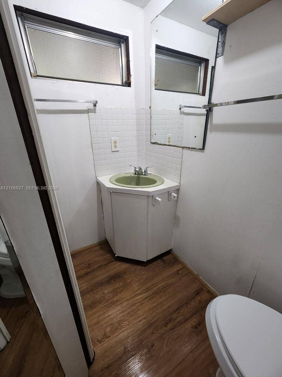 Corner vanity with an integrated basin, white tile backsplash, chrome dual-handle faucet, large wall-mounted mirror, and wood-finish flooring